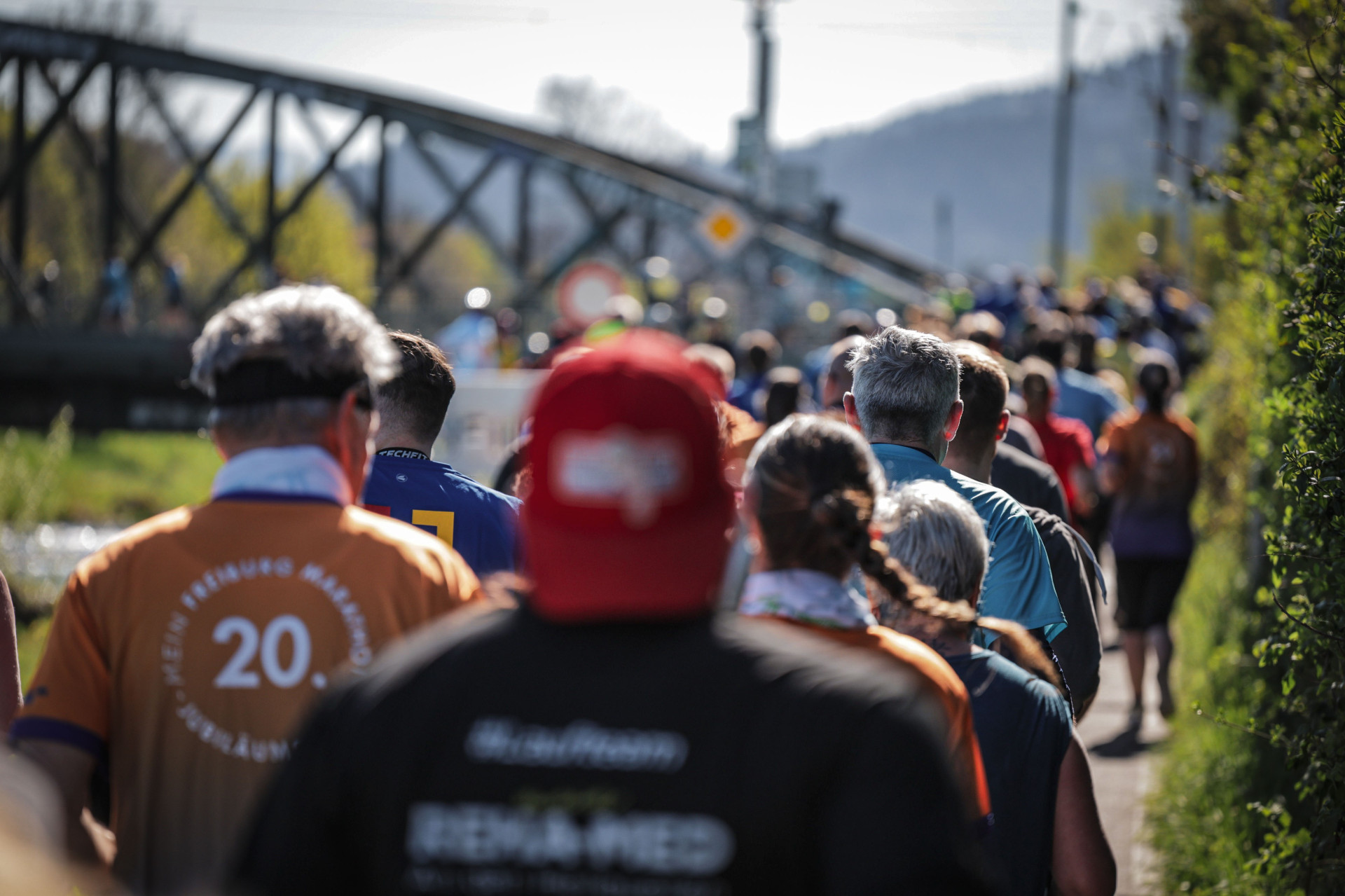 Menschenmenge bei einem Marathonlauf auf einem schmalen Weg. Die Läuferinnen und Läufer tragen bunte Sportkleidung, einige Trikots mit Aufschriften wie '20. Freiburger Marathon'. Im Hintergrund ist eine Eisenbahnbrücke und eine hügelige Landschaft zu sehen.